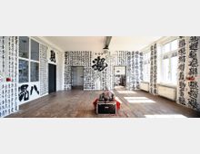 Ein heller Raum mit Holzdielenboden, weißen Wänden und japanischer oder chinesischer Kalligrafie in Schwarz, die die Wände bedeckt. Der Raum hat große Fenster, die viel Tageslicht hereinlassen, sowie drei Türen, die zu angrenzenden Bereichen führen.