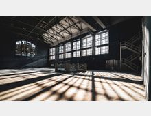 Industriehalle mit hohen Decken, großen Fensterreihen und sichtbarem Stahlträgerdach. Im Vordergrund ist eine Ziegelstruktur umgeben von Schattenwürfen, im Hintergrund eine Metalltreppe an der Seitenwand.