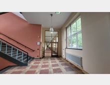 Korridor in einem öffentlichen Gebäude mit roten und beige gemusterten Fliesen, Treppe mit Glasgeländer links, großen Fenstern auf der rechten Seite und einer Durchgangstür aus Glas und Holz, die zu einem langen Flur führt. Eine Wanduhr und eine Deckenlampe sind ebenfalls sichtbar.