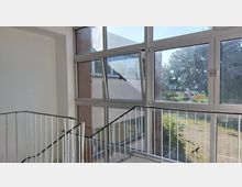 Treppenhausbereich mit weißen Wänden und einem Geländer aus weißen Metallstreben, an einer Seite große Fensterfront mit Aluminiumrahmen, die den Blick nach draußen auf Bäume, einen Parkplatz und ein Gebäude ermöglicht. Eines der Fenster ist gekippt, Licht fällt von außen ein.