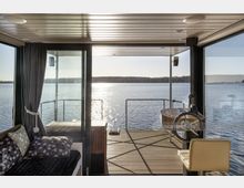 Blick aus einem modernen, verglasten Hausboot-Innenraum auf einen sonnigen See. Im Vordergrund befinden sich ein Steuerrad mit Sitz sowie ein Sofa mit Kissen, während die Holzterrasse und das Wasser im Hintergrund zu sehen sind.