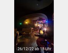 Ein gemütlicher, dimm beleuchteter Raum mit geschwungenen Decken und Vintage-Möbeln, darunter Sessel und kleine Tische. Im Vordergrund steht ein runder Tisch mit Kerzenlicht, während im Hintergrund weitere Sitzbereiche und bunte Lichtakzente sichtbar sind.