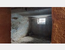 Ein leerer Kellerraum mit unverputzten Ziegelwänden, einer kleinen vergitterten Fensteröffnung, einem Betonboden und einer niedrigen Decke. Das Tageslicht fällt durch das Fenster, und der Raum wirkt verlassen und leicht verfallen.