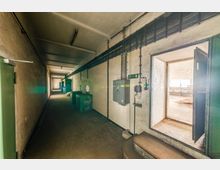 Langer, schmaler Korridor in einem industriellen oder technischen Gebäude mit blanken Wänden, freiliegenden Kabeln und einem elektrischen Schaltschrank an der rechten Seite. Rechts führt eine offene Tür in einen helleren Raum mit Fenstern und sichtbaren Rohbau-Elementen.