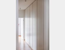 Ein schmaler, minimalistischer Flur mit einer Wand aus deckenhohen weißen Einbauschränken auf der rechten Seite und hellem Holzfußboden. Am Ende des Flurs ist eine geöffnete weiße Tür zu sehen, durch die ein weiterer Raum sichtbar wird.