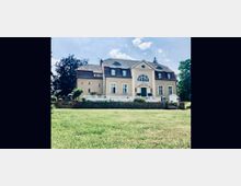 Historisches Herrenhaus mit symmetrischer Fassade, weißen Fensterrahmen und einem großen, mittig platzierten Eingangsbereich. Das Gebäude hat ein Mansarddach und ist von einer gepflegten Rasenfläche sowie Bäumen umgeben.