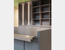 Modernes Innenraumdesign mit grauen Einbauregalen aus Holz und vertikalen Lichtelementen, kombiniert mit einem Tresen aus grauem Material und einer Edelstahl-Ablage. Im Hintergrund ist eine grün perforierte Decke sichtbar.