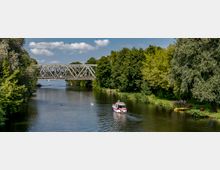 Flusslandschaft mit einer Eisenbahnbrücke im Hintergrund, umgeben von dichtem grünen Wald und Wiesen. Im Vordergrund fährt ein kleines Motorboot, daneben Schwäne und einige am Ufer liegende Kanus.