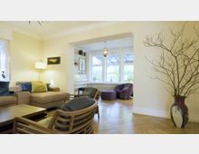 Wohnbereich mit geschmackvoller Einrichtung, darunter ein Sofa mit bunten Kissen, ein Couchtisch, moderne Holzstühle und eine Stehlampe. Im Hintergrund ein separater Sitzbereich mit großen Fenstern, die viel Tageslicht hereinlassen, sowie eine dekorative Vase mit kahlen Zweigen im Vordergrund.