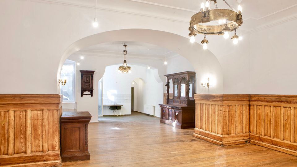 Heller Innenraum mit weißer Tonnenwölbung und großen Rundbögen, Holzdielenboden und umlaufender Holzvertäfelung. Mehrere Kronleuchter und antike Holzschränke stehen entlang der Wände, im Hintergrund sind ein Fenster und ein Durchgang zu weiteren Räumen sichtbar.