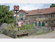 Ehemaliger Bauernhof mit Backsteingebäude, grünen Fensterläden und Ziegeldach. Im Vordergrund ein kleiner, holzumzäunter Garten mit einer Vogelscheuche in rosa Kleidung und einem aufgestellten Vogelhaus in Form eines roten Miniaturhauses.