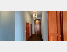 Langer, schmaler Innenflur mit hellblauen Wänden und dunklem Bodenbelag, beleuchtet durch eine moderne Deckenleuchte. Entlang des Flurs stehen mehrere braune Holztüren, am Ende eine größere Eingangstür mit verglastem Oberlicht.