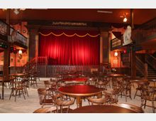 Ein Saal mit Holzmöbeln, bestehend aus runden Tischen und Stühlen, und einer Bühne mit geschlossenem roten Vorhang. Der Raum hat eine zweistöckige Galerie mit Holzgeländern, dekorative Elemente und eine rustikale Atmosphäre.