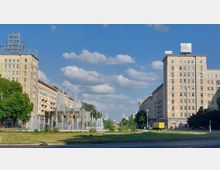 Platz mit einem Springbrunnen im Vordergrund, der von modernen Wohngebäuden im Stil des Sozialistischen Klassizismus eingerahmt wird; im Hintergrund sind zwei grüne Kirchturmspitzen sichtbar. Der Himmel ist klar mit vereinzelten Wolken, und die Szene ist von Grünflächen umgeben.