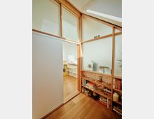 Ein modernes, lichtdurchflutetes Interieur mit Holzboden, einer Wand aus Holzrahmen und mattem Glas. Im Vordergrund steht ein Regal mit Büchern, Gläsern und Deko, während ein angrenzender Raum mit einer Schiebetür teilweise sichtbar ist.