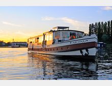 Ein stilvolles Ausflugsschiff mit dem Namen "Arcona" fährt auf einem ruhigen Fluss im Abendlicht. Im Hintergrund sind moderne Gebäude, Bäume und der Fernsehturm einer Stadt bei Sonnenuntergang zu sehen.