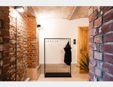 Flur mit freiliegenden Ziegelwänden, einem gewölbten Deckenbereich und einem Kleiderständer aus Metall, an dem eine schwarze Jacke hängt. Rechts befindet sich eine Holztür und eine kleine Pflanze im Vordergrund.