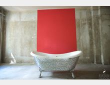 Freistehende Badewanne mit buntem Mosaikdesign vor einer Betonwand mit einem roten, rechteckigen Farbakzent. Der Raum hat einen unfertigen, industriellen Look mit sichtbaren Rohren und einer Steckdose an der Wand.