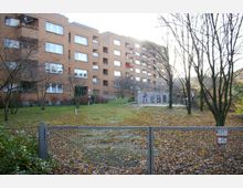 Mehrstöckiges Wohngebäude aus braunem Putz mit Balkonen, umgeben von einer Grünfläche mit kahlen Bäumen im Herbst. Im Vordergrund ein Metallzaun mit geschlossener Tür und Hinweisschildern; rechts eine bemalte Mauer mit Graffiti.