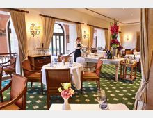 Elegantes Restaurant mit klassischer Einrichtung, weißen Tischdecken und Holzmöbeln auf einem grünen Teppich mit Blumenmuster. Große Fenster mit Vorhängen lassen natürliches Licht herein, und ein Tisch mit Blumenarrangement und Weinflaschen steht im Hintergrund.