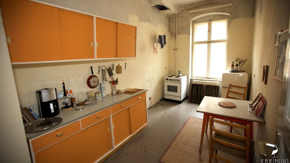 Schlichte, leicht abgenutzte Küche mit orangefarbenen Schrankfronten, langer Arbeitsplatte und frei stehendem Herd neben einem großen Fenster. Im Raum stehen ein kleiner Esstisch mit Stühlen, ein alter Kühlschrank und einfache Küchenutensilien an der Wand.
