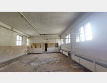 Ein leerer, verfallener Raum mit abblätterndem Putz an den Wänden, rechteckigen Fenstern mit Gitterrahmen auf der rechten Seite und einer aufgerauten Betonbodenfläche. Im Hintergrund sind kleine, abgetrennte Bereiche sowie gestapelte Holzteile zu sehen.