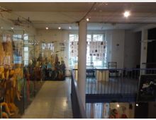 Innenraum eines Museums oder Ausstellungsraums mit Glasvitrinen voller Figuren auf der linken Seite, modernen Lichtinstallationen an der Decke, einer zentralen Säule und einem Geländer, das auf eine Etage darunter hinweist. Im Hintergrund befinden sich große Fenster und Tische mit aufgestellten Tafeln oder Plakaten.