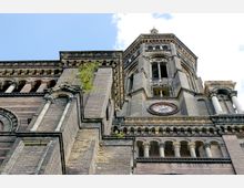 Backsteinkirche mit romanischen und neugotischen Architekturmerkmalen, darunter Rundbogenfenster, dekorative Säulen und ein Uhrenturm mit Zifferblatt. Im Hintergrund ist ein blauer Himmel mit einzelnen Wolken zu sehen.