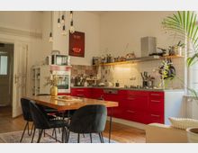 Open-plan kitchen and dining area with red lower cabinets, white tiled backsplash, and a long countertop with stove and range hood. A wooden dining table with black chairs sits under hanging pendant bulbs, with open shelving, small appliances, and potted greenery near a large window.