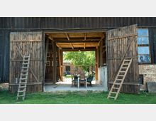 Eine offene Scheune mit hölzernen Doppeltüren, durch die ein Tisch mit Stühlen sichtbar ist. Im Hintergrund sind ein Baum sowie ein Backsteinbau zu sehen, die von Tageslicht beleuchtet werden.