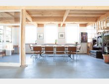 Heller Besprechungsraum mit großen Fenstern, Holzbalkendecke und einer langen Tischgruppe mit braunen Stühlen. Im Hintergrund befinden sich dekorative Pflanzen, Kunstwerke an der Wand und ein Spielautomatengehäuse.