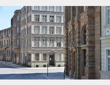 Straßenszene mit historischer Architektur, geprägt von mehrstöckigen Gebäuden mit klaren, symmetrischen Fassaden, Rechts im Bild sind verzierte Steinbögen und Gesimse zu sehen, während links ein graues Gebäude mit vielen Fenstern und einem Eingang mit Vordach dominiert. Der Boden ist gepflastert, und die Straße wirkt leer.