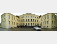 Gelbes, zweistöckiges Gebäude im neoklassizistischen Stil mit symmetrischer Fassade, hohen Fenstern und einem zentralen Eingangsbereich, der über eine breite Treppe zugänglich ist. Im Vordergrund stehen ein silbernes Auto und Gartenmöbel auf einem gepflasterten Hof.