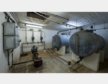 Ein technischer Raum mit zwei großen, horizontalen Metalltanks, die auf gemauerten Sockeln stehen. An den Wänden verlaufen Rohrleitungen mit Ventilen, daneben befinden sich elektrische Schaltkästen.