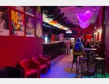 Eine Bar mit roter Wandgestaltung und schwarzen Theken, beleuchtet durch farbige Lichter. Im Raum hängen dekorative Regenschirme von der Decke, und Gäste sitzen an Tischen oder an der Bar.