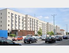 Mehrstöckiges Wohngebäude mit heller Fassade und zahlreichen kleinen Balkonen, im Vordergrund eine stark befahrene Straße mit Autos und einem Lieferwagen, daneben eine kleine Grünfläche und vereinzelte Bäume.