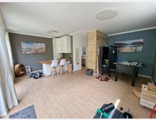 Ein multifunktionaler Wohnraum mit Holzfußboden, einer kleinen Küche mit weißer Ausstattung und einer Bartheke mit zwei Hockern. Im Hintergrund befinden sich eine Holzwand, ein Kickertisch und eine blaue Wand mit großformatigem Landschaftsbild; seitlich stehen Fitnessgeräte und ein Regal.