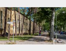 Wohnstraße mit gelb gestrichenen Mehrfamilienhäusern, die von hohen Bäumen flankiert sind. Ein gepflasterter Gehweg verläuft entlang der Gebäude, daneben eine Straße mit parkenden Autos und vereinzelt gehenden Personen.