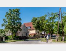 Straßenszene in einer Wohngegend mit freistehenden Häusern, umgeben von Bäumen und Gärten. Im Vordergrund fährt eine Person mit dem Fahrrad auf einem gepflasterten Weg, während Holzmasten und ein Verkehrsschild sichtbar sind.