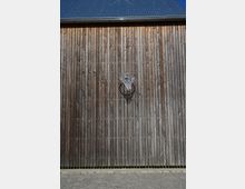 Holzwand eines Gebäudes mit vertikalen Holzlatten, darauf ein Basketballkorb montiert; im oberen Bereich ist ein dunkles Blechdach mit Dachrinne sichtbar. Darunter befindet sich ein Kiesboden.