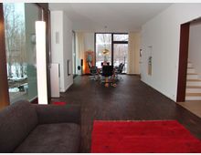 Modernes Wohnzimmer mit dunklem Holzfußboden, roten Teppichen und großen Fenstern, die einen Blick auf eine schneebedeckte Außenszenerie bieten. Im Hintergrund befindet sich ein Esstisch mit Stühlen, daneben eine Treppe und helle Wände.