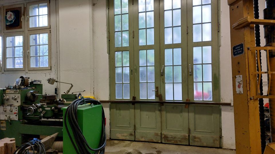 Ein Werkstattraum mit großen, grün lackierten Doppeltüren und Glasfenstern, die von Holzrahmen umgeben sind. Im Vordergrund steht eine grüne Drehmaschine, daneben Kabel und Maschinenteile; rechts ist ein Teil einer gelben Hebevorrichtung sichtbar.