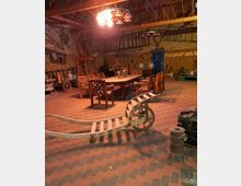 Ein rustikaler Innenraum einer Scheune mit roten und grauen Ziegelböden. Im Mittelpunkt stehen ein hölzerner Esstisch mit passenden Stühlen und eine alte Schubkarre; an den Wänden lehnen landwirtschaftliche Werkzeuge und es hängen rustikale Dekorationselemente.