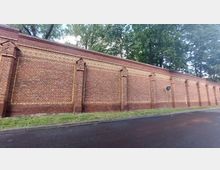 Lange Mauer aus roten Ziegelsteinen mit gelben Akzenten und vertikalen Stützpfeilern, am unteren Rand mit Gras und einem angrenzenden asphaltieren Gehweg; im Hintergrund sind große Bäume sichtbar.