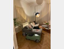 Wohnzimmer mit hellen Wänden, einem hölzernen Fischgrätboden und einer grauen Eckcouch, auf der Kissen liegen. Links steht eine Stehlampe mit rundem Lampenschirm, rechts befinden sich zwei runde Tische mit Blumenvasen, und im Vordergrund steht ein grünes Spielzeugauto.