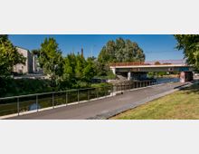 Ein asphaltierter Weg mit Geländer verläuft entlang eines kleinen Kanals, der von einer roten Brücke überspannt wird. Im Hintergrund sind Bäume, ein Gebäude und eine mit Graffiti verzierte Mauer zu sehen.