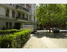 Wohnstraße in einer städtischen Umgebung mit mehrstöckigem Gebäude im historischen Stil, großen Fenstern und kleinen Balkonen. Im Vordergrund befindet sich eine Hecke neben einem Gehweg, während ein Baum, ein parkender Lieferwagen und ein Metallcontainer den Platz beschatten.