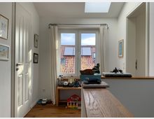 Heller Flur in einem Wohnhaus mit weißen Wänden und Holzboden. Ein großes Fenster mit Blick auf ein Nachbarhaus mit roten Dachziegeln dominiert den Raum; darunter steht ein kleiner Tisch mit Spielzeugautos.