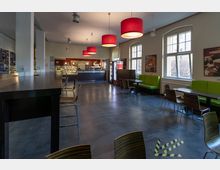 Ein moderner Cafeteria-Raum mit hohen Tischen, Stühlen und Sitzbänken mit grünem Polster entlang der Fenster. Im Hintergrund befindet sich eine Selbstbedienungstheke mit warmem Licht und Regalen, sowie rote Hängelampen an der Decke.