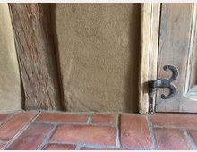 Nahaufnahme eines Wandabschnitts mit einer Lehmputz-Oberfläche, einem vertikalen Holzbalken und einer hölzernen Tür mit dekorativem Metallscharnier. Der Boden besteht aus roten Terrakottafliesen mit Fugen.